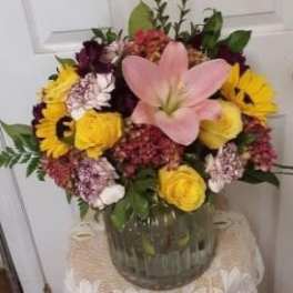 Mixed bouquet with a pink lily, yellow roses, and purple flowers in a glass vase