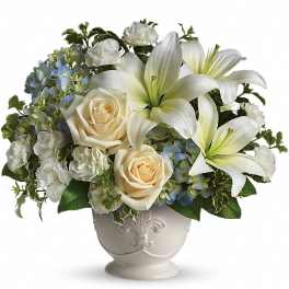 Low arrangement of white lilies, cream roses, white carnations and blue hydrangeas in a white ceramic footed bowl