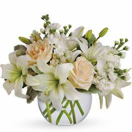 Low arrangement of white lilies, cream roses, and white flowers in a round clear glass vase
