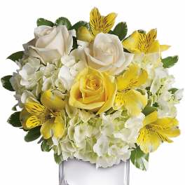 Short arrangement of white hydrangeas, yellow and white roses, and yellow alstroemeria in a glass cube vase
