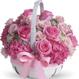 Round basket arrangement of pink roses and hydrangeas with a white handle and pink ribbon bow.