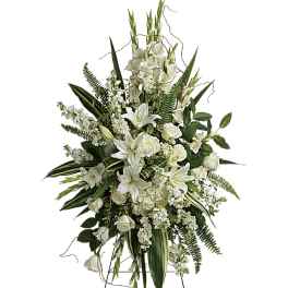 Standing spray of white lilies, roses, and other blooms with tall green foliage on a metal easel.