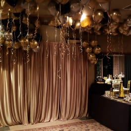 Gold and black balloons with curling ribbons over a draped party backdrop