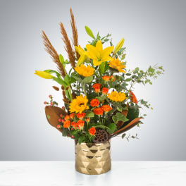 Tall yellow lilies, sunflowers, and orange roses arranged in a gold geometric vase