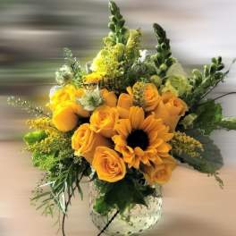 Yellow roses and a sunflower arranged with accent blooms in a clear textured glass vase