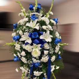 Tall standing spray of blue roses, white lilies, and white mums with blue ribbon on an easel.