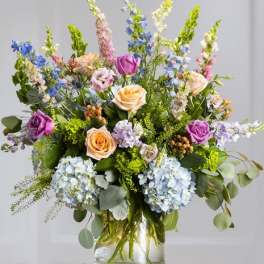 Mixed arrangement of lavender and peach roses, blue hydrangeas, and tall delphinium in a clear glass vase