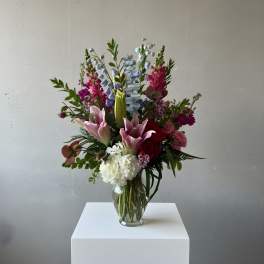 Tall mixed arrangement with pink lilies, red roses, white hydrangea, and blue delphinium in a clear glass vase.