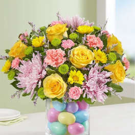 Bouquet of yellow roses, pink chrysanthemums, and carnations in a glass vase filled with pastel eggs.