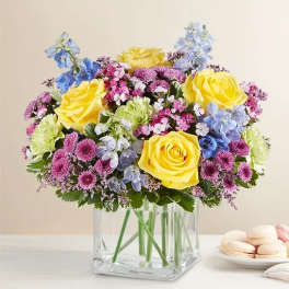 Bouquet of yellow roses, blue and pink flowers in a clear square vase