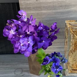 Purple orchid arrangement in a brown vase beside a small blue flower bouquet in a glass bottle
