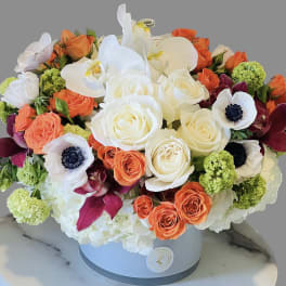 Round bouquet of white orchids, roses, and orange blooms in a hatbox.