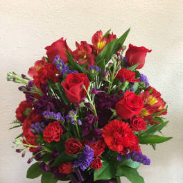 Tall arrangement of red roses and carnations with purple flowers in a glass vase with a purple ribbon.