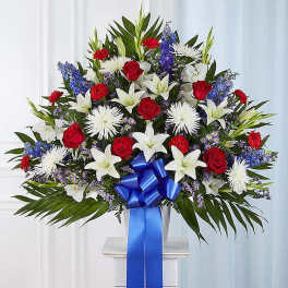 Tall red, white, and blue sympathy arrangement with roses, lilies, mums, and a large blue ribbon bow