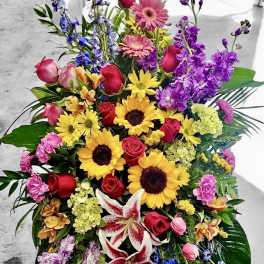 Tall standing spray of mixed bright flowers including sunflowers, roses, lilies, and gerbera daisies with palm fronds