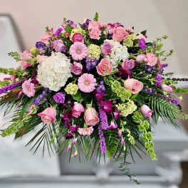 Pink and purple casket spray with roses, gerbera daisies, orchids, and hydrangeas on a white casket.