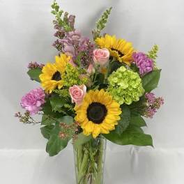 Tall mixed bouquet with sunflowers, pink roses, purple blooms, and green hydrangea in a clear glass vase