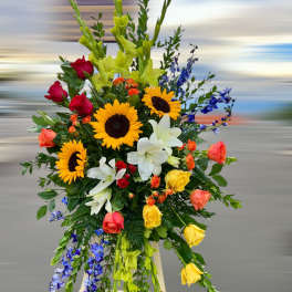 Tall standing spray of sunflowers, roses, lilies, and blue delphinium on a wooden easel