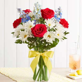 Bouquet of red roses, white daisies, and blue flowers in a glass vase with a yellow ribbon