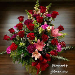 Tall arrangement of red roses, pink lilies, and carnations in a clear vase with a red bow