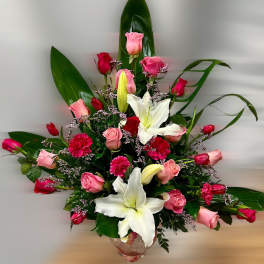 Tall arrangement of pink and red roses, white lilies, and bright pink carnations in a clear glass goblet vase