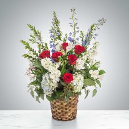 Tall red, white, and blue flower arrangement with roses and delphinium in a woven basket