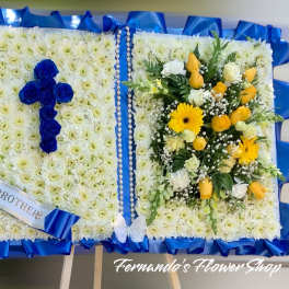 Standing open-book funeral arrangement with blue rose cross and yellow and white flowers.