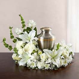 White lily and rose cremation wreath surrounding a silver urn on a dark wood table