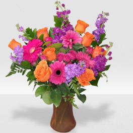 Bright arrangement of orange roses, hot pink gerbera daisies, and purple flowers in a brown glass vase.