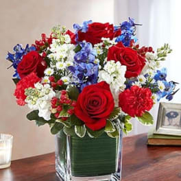 Bouquet of red roses, blue flowers, and white daisies in a square glass vase