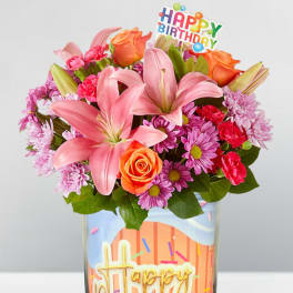 Birthday bouquet with pink lilies, orange roses, and purple daisies in a glass vase