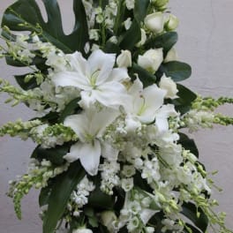 Tall standing spray of white lilies, roses, and other white flowers with large dark tropical leaves.