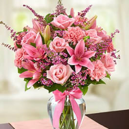 Pink roses, lilies, carnations, and filler flowers in a clear glass vase with a pink ribbon