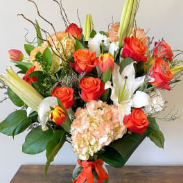 Tall mixed arrangement of orange roses, tulips, white lilies, and peach hydrangeas in a clear vase with orange ribbon