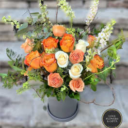 Arrangement of orange and cream roses with tall white blooms in a gray cylinder vase