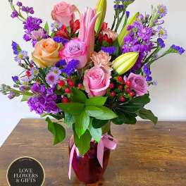 Mixed bouquet of pink roses, lilies, and purple flowers in a red glass vase with pink ribbon.