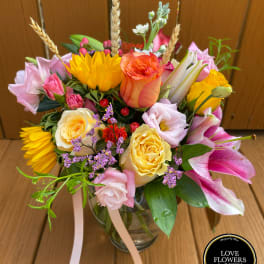 Colorful bouquet of sunflowers, roses, lilies, and mixed blooms in a clear glass vase with ribbons.