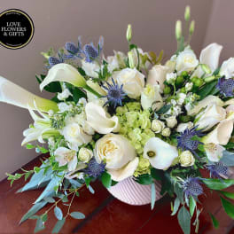 Low white and green arrangement of roses, calla lilies, and blue thistle in a white ribbed vase