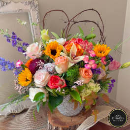 Colorful arrangement of sunflowers, roses, and lilies in a white ceramic pot with twig accents.