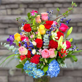 Tall mixed bouquet of multicolored roses, lilies, irises, and blue hydrangeas in a glass vase