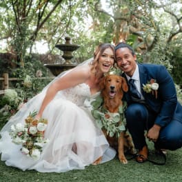 Bride and groom posing with a dog, each holding a floral bouquet