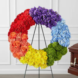 Rainbow floral wreath on a black stand beside a casket