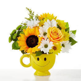 Cheerful bouquet of sunflowers, white daisies, and a yellow rose in a yellow smiley-face mug.