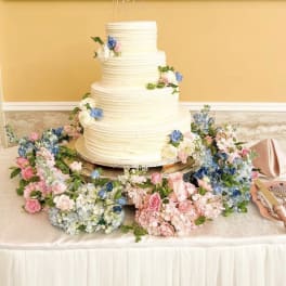 Four-tier white wedding cake surrounded by pastel floral arrangements