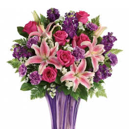 Pink lilies, roses, and purple flowers arranged in a tall purple glass vase