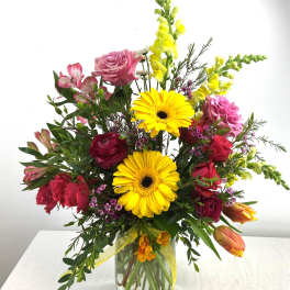 Tall mixed bouquet of yellow gerbera daisies, pink roses, and bright blooms in a clear glass vase.