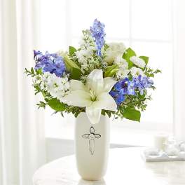 White lilies and blue flowers arranged in a cream vase with a cross emblem