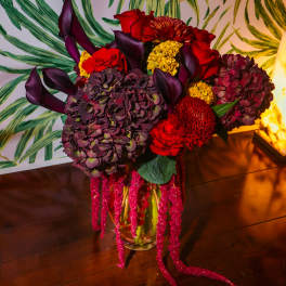 Jewel-toned arrangement of red roses, dark calla lilies, and hydrangeas with trailing pink accents in a glass vase.