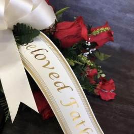 Sympathy spray with red flowers and a white ribbon reading Beloved Father, accented by a large ivory bow.