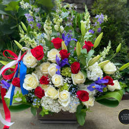 Tall red, white, and blue arrangement with roses, lilies, irises, and a patriotic ribbon in a rectangular container